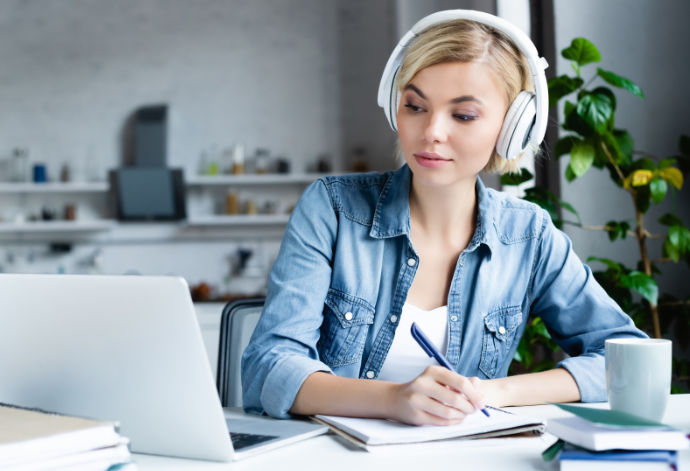 Young blond woman with headphones, holding a pen over a jotting pad and looking right at her laptop