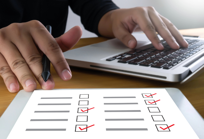 Person's left hand on laptop keyboard, right hand holding a pen and touching a checklist